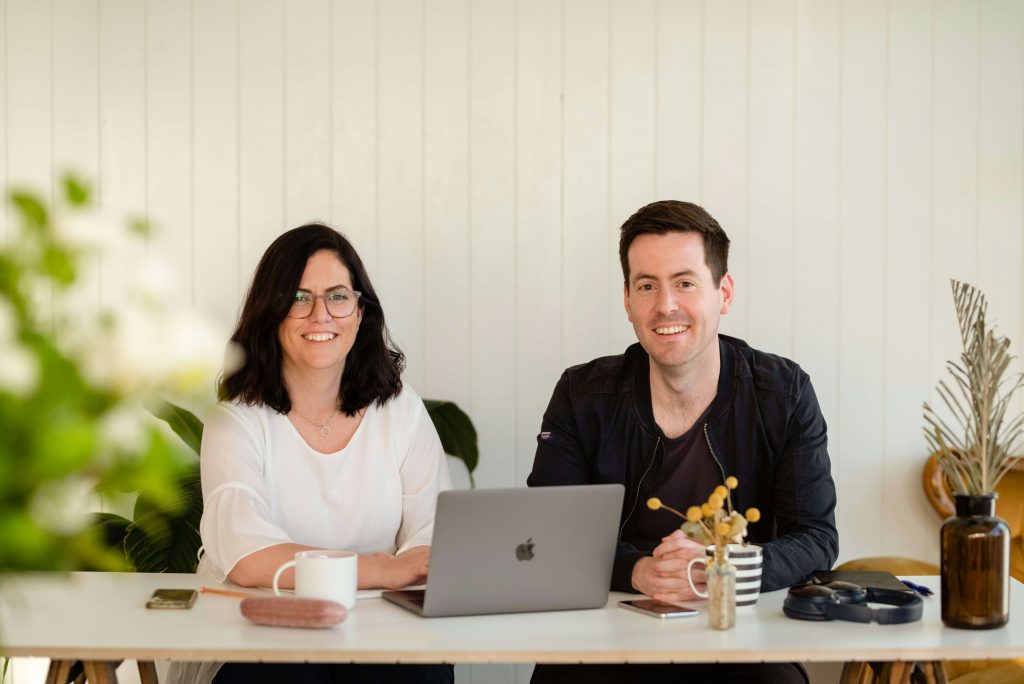 1 Mann und eine Frau sehen lächelnd in die Kamera, vor ihnen steht ein aufgeklapptes Macbook
