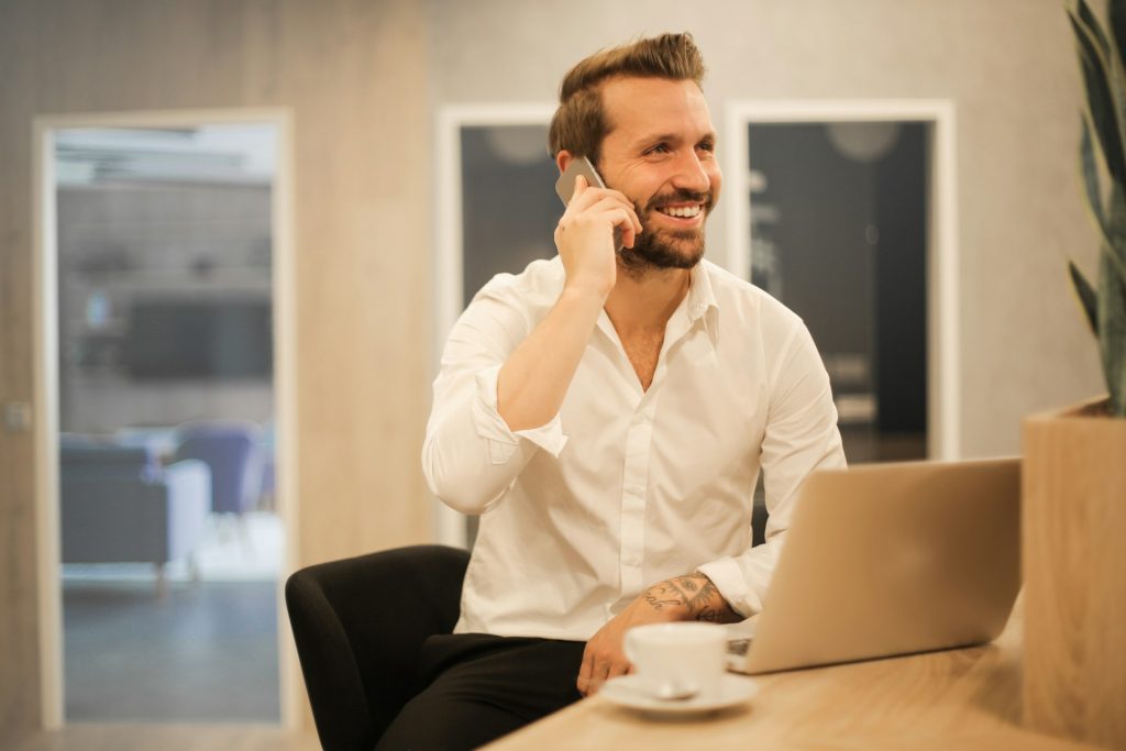 Mann lacht und hat Handy am Ohr, sittz am Schreibtisch vor Notebook und Tasse Kaffee